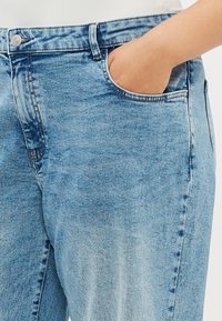 Personne portant un jean denim bleu clair, la main droite placée dans la poche avant sur fond blanc.