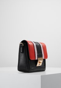 Borsa in pelle nera con accenti rossi e rosa chiaro, chiusura rettangolare in tono oro, appoggiata su un piedistallo bianco contro uno sfondo grigio.