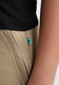 Kakhibukser med en tekstureret finish og en sort skjorte. Har et lille turkis mærke på bukserne nær sømmen.