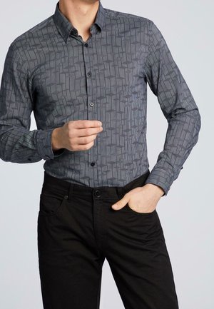 Mann trägt ein eng anliegendes, langärmeliges Hemd mit geometrischem Muster und schwarze Hose, mit einer Hand in der Tasche.