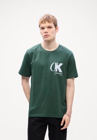 Tamnozelena pamučna majica s bijelim logotipom "CK" i natpisom "Calvin Klein EST 1968" na prednjoj strani. Okrugli izrez i kratke rukave.