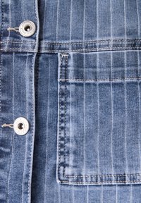 Denimkangas sinisillä raidoilla, jonka metalli-nappikoristeet ja vahvistetut ompeleet. Mukana etutasku, jossa näkyvät sauman yksityiskohdat.