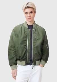 Green bomber jacket with a smooth texture, ribbed cuffs, snap buttons, side pockets, and a zippered pocket on the sleeve.