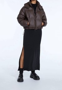 Giacca puffer marrone con zip frontale, polsini elastici e fondo con coulisse, abbinata a un vestito lungo nero aderente con una fessura laterale.
