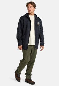 Marine blauwe hoodie met rits, trekkoord, voorzakken en logo; gedragen over een licht T-shirt en gecombineerd met olijfgroene broek en bruine wandelschoenen.