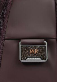 Sezione di borsa color borgogna scuro con un'etichetta rettangolare in pelle con le iniziali "M.P." in rilievo e una cornice metallica argentata. Dettaglio della superficie testurizzata visibile.