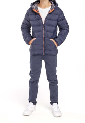 Chaqueta acolchada azul marino con capucha, con cremallera frontal, dos bolsillos y detalles en naranja contrastante. Lleva pantalones grises y zapatillas blancas.