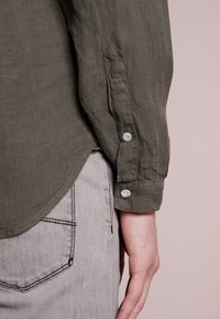 Olivově zelená lněná košile s knoflíkovými manžetami, zdobená světlomodrými knoflíky a zakřiveným lemem, ve spojení se šedými džíny.