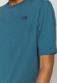 Tyrkysové tričko s krátkým rukávem vyrobené z bavlny. Na hrudi má malé černé logo. Klasický kulatý výstřih s hladkou texturou.