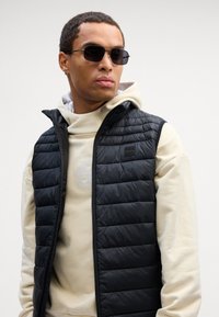 Μαύρο αμάνικο μπουφάν με φερμουάρ μπροστά, πλισέ υφή και ψηλό λαιμό, το οποίο είναι στρωμένο πάνω από μία ανοιχτόχρωμη μπλούζα με κουκούλα.