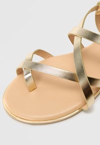Kulta metallinen nauhakenkä litteällä pohjalla, jossa on sileä beige sisäpohja ja pyöreä kärki, korostettuna ohuella kultaisella reunalla.