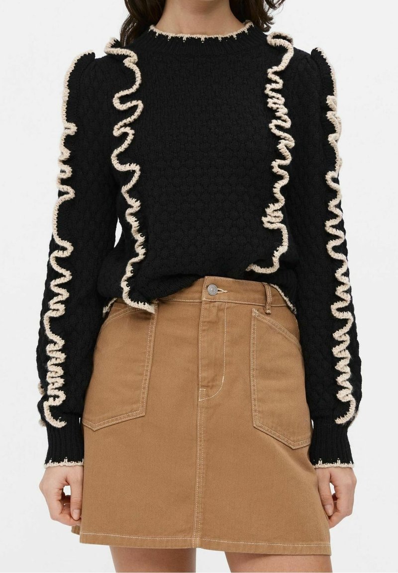 Femme portant un pull noir texturé avec un bord volanté crème et une jupe marron taille haute en ligne A avec poches plaquées à l'avant.