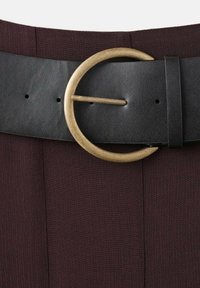 Cintura in pelle nera con una grande fibbia circolare in metallo dorato, caratterizzata da una superficie testurizzata e uno sfondo in tessuto bordeaux scuro.