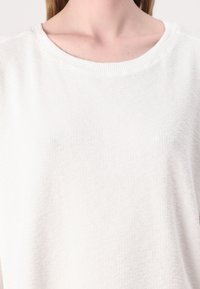 Hvit teksturert kortermet skjorte med rund hals brukt av en person vist fra nakken til øvre del av brystet.