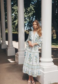 White dress with blue floral patterns and green accents, long sleeves, tiered skirt, and a waist tie. Worn with white sandals.