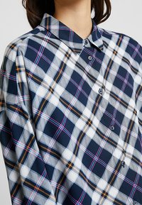Camicia a quadri blu navy e bianca con linee contrastanti in rosa e arancione, dettagli con bottoni e colletto piegato; realizzata in un tessuto testurizzato.