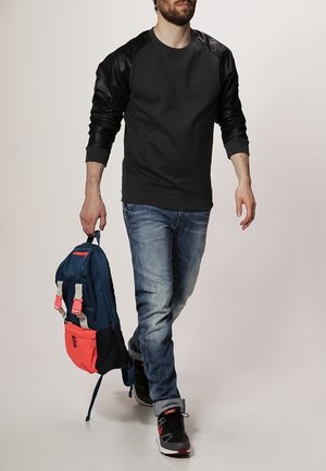 Sudadera de cuello redondo gris oscuro con mangas de cuero negro, combinada con jeans azules. Lleva una mochila azul y rosa con acentos blancos.