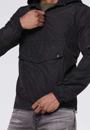 Homme portant une veste coupe-vent noire à capuche avec une fermeture éclair et une poche à l'avant, ajustant la fermeture éclair d'une main.