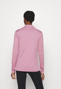 Camicia a maniche lunghe di colore rosa chiaro, realizzata in tessuto morbido con colletto, caratterizzata da una superficie liscia e un design aderente, indossata con leggings neri.