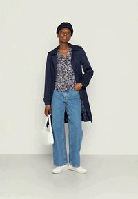 Mulher vestindo um casaco azul-marinho, blusa floral, jeans azul-claro, tênis brancos e boné azul-marinho, segurando uma bolsa branca em frente a um fundo simples.