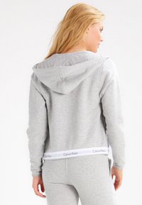 Hanorac gri cu glugă, dintr-un material moale, prezentând un band cu logo-ul alb al Calvin Klein la talie și un croi lejer, casual.