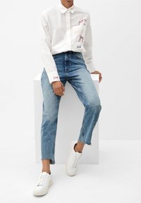 Camisa blanca de botones con texto bordado y bolsillo, combinada con jeans de mezclilla azul con dobladillo sin acabar, y zapatillas blancas.