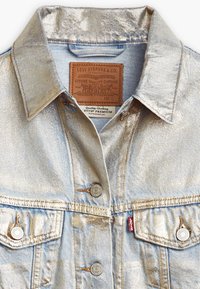 Ljusblå denimjacka med metallisk lyster, klassisk krage, knäppning framtill, två bröstfickor och en läderlogotyp på nacken.