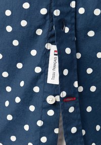 Camicia button-up blu navy con pois bianchi, etichetta in tessuto con "Tissu Français", cuciture di accento rosse e dettaglio di un singolo bottone bianco.