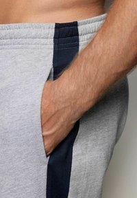 La mano derecha colocada dentro del bolsillo lateral de unos pantalones de chándal grises con una raya vertical oscura cerca de la cinturilla.