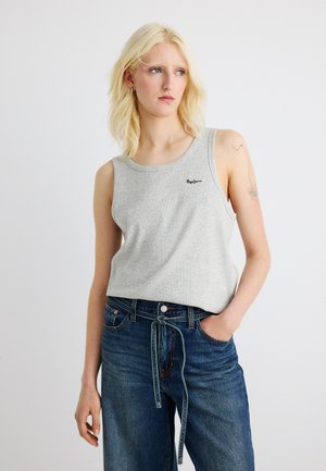 Donna bionda con tatuaggio sul braccio superiore che indossa una canotta grigia e jeans blu a vita alta con cintura annodata, in piedi su sfondo bianco.