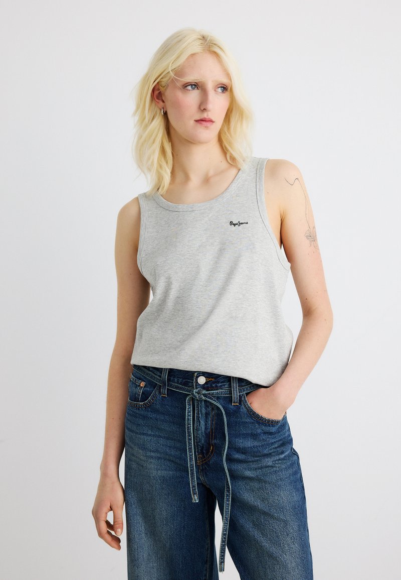 Ξανθιά γυναίκα με τατουάζ στον πάνω βραχίονα, φορά γκρι αμάνικη μπλούζα και μπλε τζιν ψηλόμεση με δεμένη ζώνη, στέκεται σε λευκό φόντο.