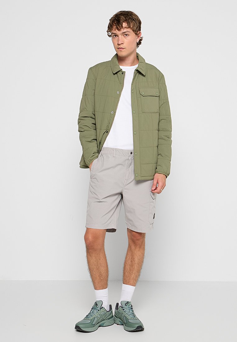 Lätt grön vadderad jacka över en vit t-shirt, ihop med beige shorts och olivgröna sneakers. Bakgrunden är helt vit.