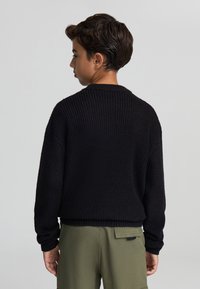 Czarny sweter dzianinowy o ryflowanej fakturze, okrągłym dekolcie i opadających ramionach, zestawiony z oliwkowymi spodniami.