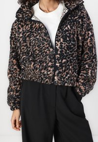 Chaqueta recortada de tejido suave con estampado de leopardo en tonos negros, beiges y rosas. Presenta un cuello alto y cierre de cremallera negro.