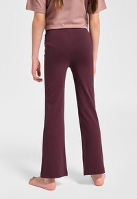 Osoba stojąca boso, ubraną w ciemnofioletowe dopasowane spodnie i krótkim rękawem w kolorze przygaszonego różu na jednolitym tle.