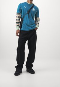 Maglia sportiva turchese con maniche lunghe a righe, pantaloni neri larghi e sneakers nere. Presenta dettagli del logo e cuciture a contrasto.