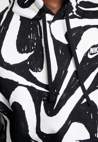 Svart och vit huvtröja med abstrakta penseldragmönster, dragsko och en liten broderad logotyp på bröstet. Material av bomullsblandning.