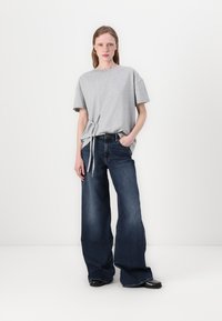 Gray cotton oversized t-shirt with side tie detail paired with wide-leg dark blue denim jeans. Black shoes complete the outfit.