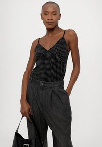 Camiseta de tirantes finos negra con lunares, combinada con pantalones grises de tiro alto. La modelo sostiene un bolso negro brillante. Textura de tela suave.