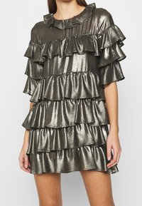 Puku metallinen kiiltävä mekko, jossa on useita röyhelökerroksia, lyhyet hihat ja pyöreä pääntie. Väri on tumma hopea ja pinta on sileä.