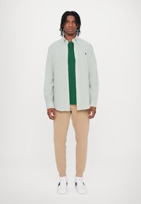 Cămașă cu mâneci lungi în carouri verzi și albe, purtată peste un tricou verde, asociată cu pantaloni jogger bej și adidași albi cu accente bleumarin.