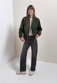 Olivgrön bomberjacka med ribbad krage, tillsammans med grå raka jeans och vita sneakers med blå accenter.