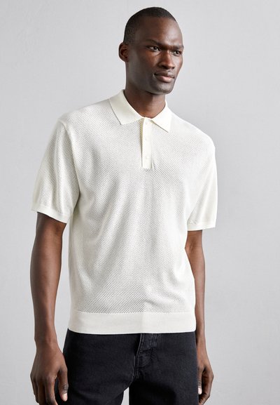 Polo à manches courtes en crème clair, avec un motif en maille texturée, un col classique et une patte de boutonnage à trois boutons. Associé à un jean noir.