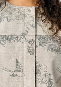 Blouse en tissu gris avec un imprimé floral et d'oiseaux, doté d'une finition texturée, d'un col rond et de coutures centrales décoratives.