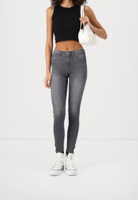 Grijze skinny jeans met een hoge taille en stretchstof, gecombineerd met een zwart cropped tanktop en witte sneakers, met een kleine witte tas in de hand.