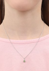 Silverfärgad kedja med ett litet, grönt ädelsten-hänge i form av en blomma, buren över en rosa skjorta. Enkel design och delikat textur.