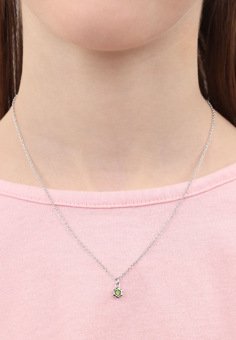 Silverfärgad kedja med ett litet, grönt ädelsten-hänge i form av en blomma, buren över en rosa skjorta. Enkel design och delikat textur.