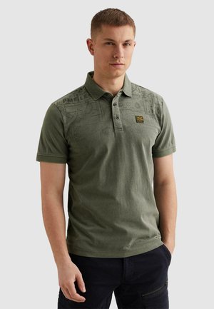 Muškarac u maslinasto zelenoj polo majici s printom i crnim hlačama, stoji s jednom rukom u džepu na svijetlosivoj pozadini.