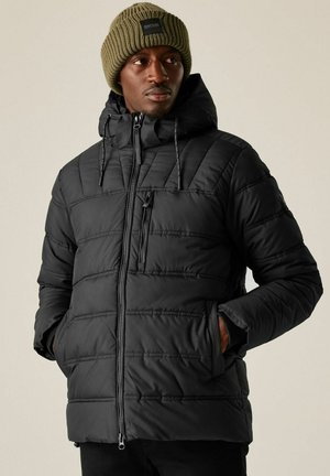 Mężczyzna ubrany w czarną pikowaną kurtkę z kapturem i oliwkowo-zieloną dzianinową czapkę, ręce w kieszeniach, patrzący na bok na jednolitym tle.