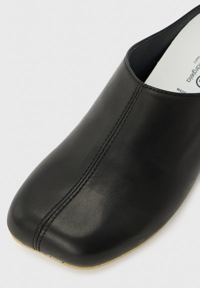 Μαύρο δερμάτινο παπούτσι τύπου slip-on με τετράγωνη μύτη και κεντρική ραφή, εμφανιζόμενο με μέρος της λευκής εσωτερικής σόλας και της μπεζ άκρης της σόλας.